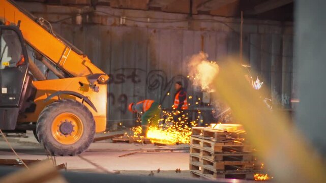 A subcontractor, wearing a helmet and safety gear, is cutting metal structures with an angle grinder in a construction site. Sparks fly as work continues indoors under artificial lighting