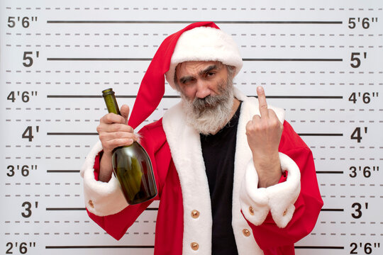 Bad Santa Claus with an offensive obscene gesture drunkard with bottle of wine, Police Mugshot Board backdrop of a ruler with height. Natural beard, red hat, suit