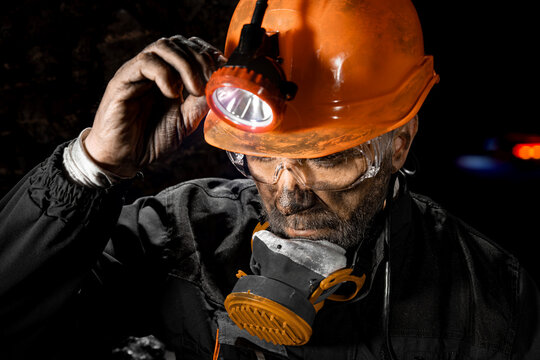 Coal miner with a dirty face in black dust, wearing a protective helmet with a flashlight, filtering respirator and work clothes for working in mine. Hard work underground in a coal mine, digging site