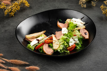 Fresh duck salad with pear slices, tomatoes, leafy greens and creamy cheese on a dark plate