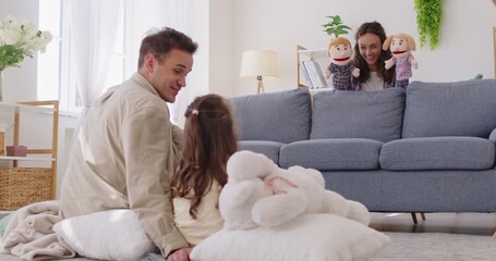 Happy playful family at home play puppet theater show with child girl. Parents and daughter animate plush characters on sofa, laughing together indoors. Family bonding and leisure concept.