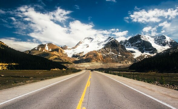 An empty asphalt road stretches towards a majestic snow-capped mountain range under a blue sky - Powered by Adobe