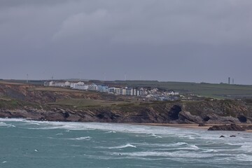 An overcast landscape shows a distant shoreline. White buildings line the coast, with green hills beyond a sandy beach and rocky cliffs