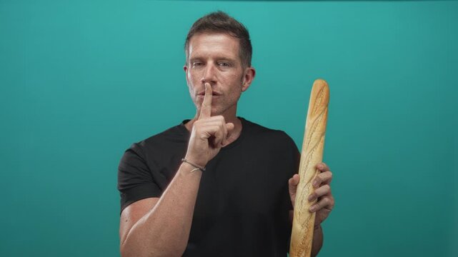 Man holding baguette and finger to lips for silence in teal studio with casual black t shirt and bracelet visible; silence.