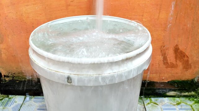 Close-up of a plastic bucket overflowing as water continues to pour from above. Strong bubbling and splashing highlight pressure, waste, and everyday water usage in a simple household setting.