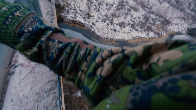 A man walks dangerously along the edge of the bridge in winter, holding onto the railing. A risky move. A guy climbs over a frozen river. From the first person.