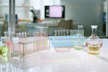 Laboratory glassware including test tubes, beakers, flasks, petri dish and microplate containing various colored liquids arranged on laboratory table with blurred scientific equipment in background