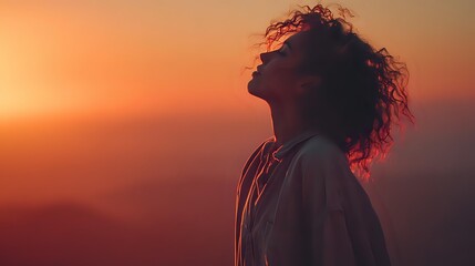 Silhouette of woman with curly hair against dramatic sunset sky, enjoying peaceful moment on mountain overlook.