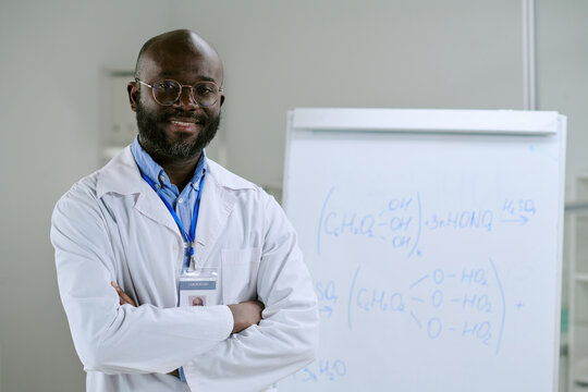 Portrait of middle aged Black man wearing lab coat and identification badge standing with arms crossed in laboratory near whiteboard displaying chemical formulas - Powered by Adobe