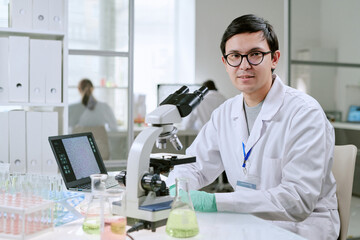 Portrait of young adult man wearing eyeglasses sitting at laboratory workstation operating microscope with laptop and glassware visible