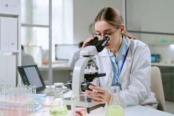Young adult Caucasian woman examining samples through microscope in laboratory, conducting medical research surrounded by glassware