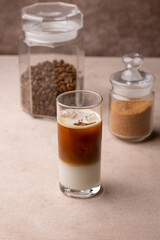 Coffee with coconut milk and ice cubes in a tall glass. Traditional non-alcoholic cold drink. In the background, a jar of coffee and a jar of sugar. Selective focus, close-up.