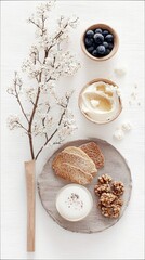 A top-down view of a healthy breakfast spread on a white wooden surface. Includes a wooden vase with white blossoms, a bowl of blueberries, a bowl of yogurt wit