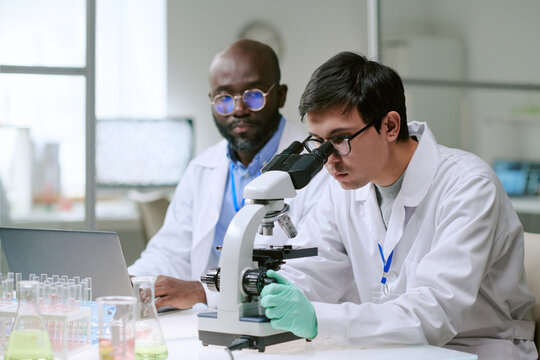 Caucasian man examining sample with microscope while Black man observing and working on laptop in laboratory setting with scientific glassware visible