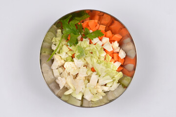 An overhead studio shot shows a stainless steel bowl filled with diced carrots, chopped cabbage, and cauliflower pieces with parsley on a white background