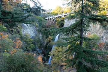 Tbilisi, Georgia - November 03, 2025: Majestic waterfall cascading down rocky cliff surrounded by vibrant autumn foliage and a picturesque bridge in a serene natural landscape