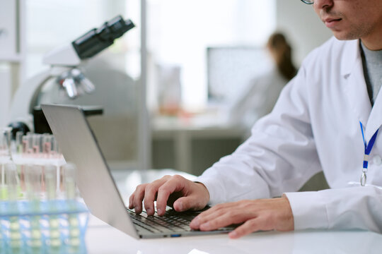 Young adult Caucasian man wearing lab coat working on laptop in laboratory, hands typing on keyboard near test tubes and microscope, female scientist blurred in background