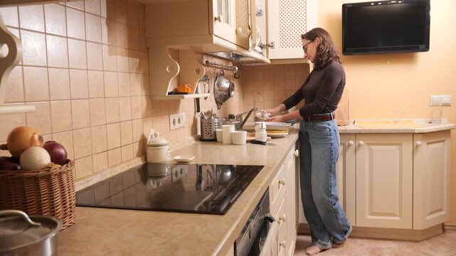 Woman standing at kitchen sink washing white ceramic dishes with a sponge and suds, focused on daily chores in a cozy, modern home environment while wearing casual denim and glasses