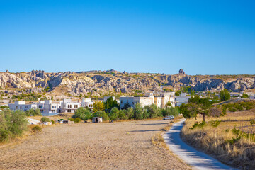 A picturesque scene of a small village in Cappadocia, Turkey, surrounded by dramatic rock formations and soft, eroded hills. The warm afternoon light highlights the white stone houses, green trees