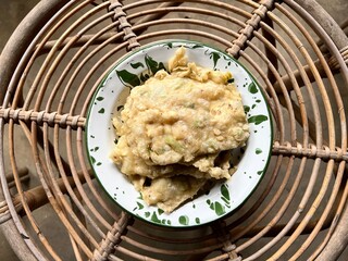 Tempe Mendoan, an Indonesian traditional snack made from tempe or tempeh (fermented soybeans) coated in flour and spring onions and fried half-cooked, on a white enamel plate, placed on a rattan table