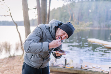 Male enjoying outdoor meal by serene lake, wearing gray jacket and black beanie, surrounded by nature, showcasing a moment of relaxation and connection with the environment