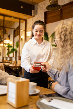 Vertical photo of a young girl with Down syndrome working in a modern cafe wearing a white uniform.The girl is taking a payment from a customer using a card reader.Concept of trisomy 21.
