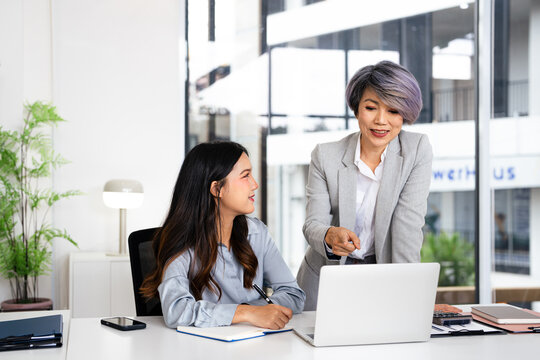 Professional mature business woman manager explaining solutions helping young employee at work. Busy business people using laptop talking in office.