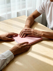 Two men's hands rest together on a pink book in a gesture of support and trust. Partnership and connection concept