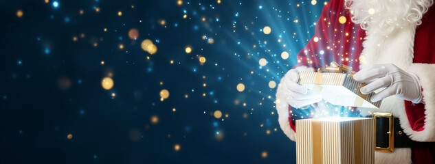 Santa Claus hands opening gift box with magical beam and stars. Blue panoramic background with room for text