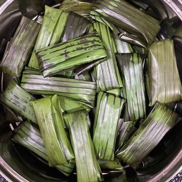 A close up top view of traditional Malaysian lepat pisang wrapped in fresh green banana leaves, arranged neatly in a stainless steel container. Ideal for themes related to Asia traditional cuisine