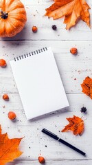 A flat lay composition featuring a whole pumpkin, an open blank notebook, a black pen, and scattered autumn leaves and nuts on a rustic white wooden surface. Th