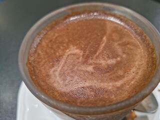 Close-up, top-down view of a glass mug filled with hot chocolate or cocoa, featuring a frothy, rich brown surface with a subtle swirl.