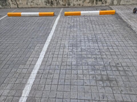 An empty outdoor parking space defined by a white painted line and two concrete wheel stops painted white and yellow/orange, on a gray stamped concrete or block pavement.