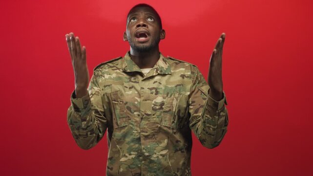 Soldier man in camouflage uniform raises hands and looks upward against a solid red studio backdrop; hope.