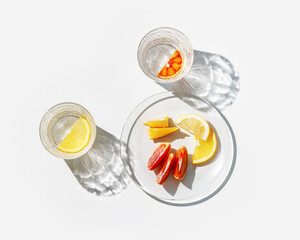 Clear Glasses and plate as summer still life with shadow and sunlight reflection on white table, close up glass cups of infused fruit water, slises of lemon, orange, tangerine, healthy summer drink