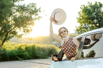 Joyful woman in vintage polka dot dress waving her straw hat from car window. Retro summer travel mood, freedom, happiness, and vacation adventure concept