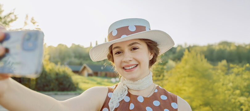 Smiling woman in vintage polka dot dress and straw hat taking selfie with smartphone against nature background. Summer lifestyle moment, freedom, joy, and retro charm.