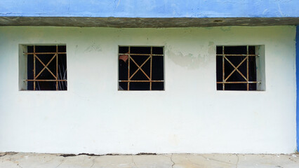 Three small, square windows with rusted iron security bars (grilles) set symmetrically in a distressed white exterior wall with a blue trim. Concept of isolation, decay, and confinement.