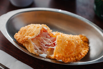 Close-up photo of a deep-fried empanada, split open to show the gooey, stretching melted cheese and sliced ham filling. The golden-brown, crispy pastry sits in a stainless steel serving dish.