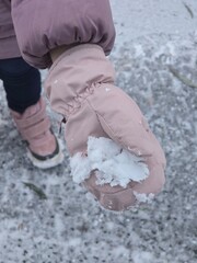 A close-up of a child's hand wearing a pink, insulated mitten and holding a small clump of white snow, with winter boots visible in the background.