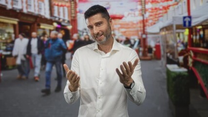 Man smiling with open hands gesture in crowded street market during daytime, wearing white shirt; warmth and approachability.