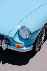 Close-up of the front corner of a light blue vintage car, showing its round headlight, chrome bumper, indicator light, and classic wire wheel.