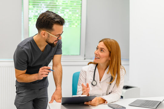Male physician reviewing diagnostic reports with confident female colleague at desk in modern hospital - Powered by Adobe