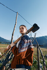 Naklejka premium Woman with axe standing by wooden fence in mountains field, wearing patterned sweater on a rural farm. Portrait outdoors with rustic wood posts, clear blue sky and natural sunlight.