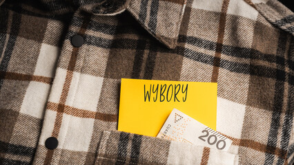 Yellow card with a handwritten inscription "Wybory", protruding from a brown plaid shirt, next to Polish banknotes PLN (selective focus)