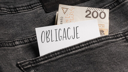 White card with a handwritten inscription "Obligacje", inserted into the pocket of gray pants jeasnow, next to Polish banknotes PLN (selective focus)