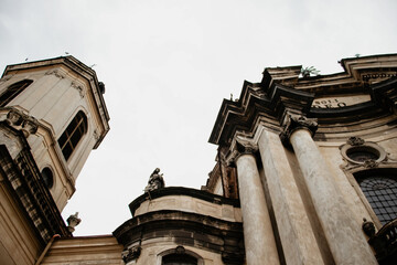 Low angle view of grand Baroque cathedral architecture featuring massive stone columns, ornate sculptures, and a high tower against a pale overcast sky in an old European city.