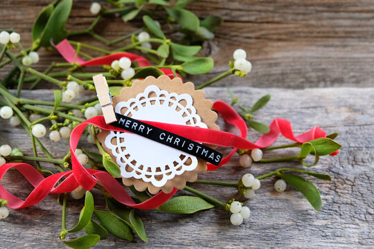 "Merry Christmas" greeting paper card, red ribbon and Mistletoe plant green branches close up on wooden background. decor for Christmas, Advent holidays, festive winter season. top view