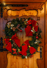 A vertical shot of a Christmas wreath with a bright red bow on a rustic wooden door. Perfect for holiday promotions, seasonal greetings, home decor visuals, and classic Christmas-themed designs.