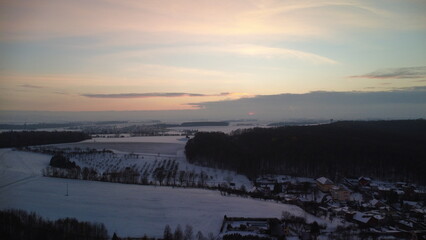 Sunset over a snow-covered Czech landscape, featuring rolling fields, trees, and a serene winter atmosphere. The warm colors of the setting sun contrast beautifully with the fresh snow.
